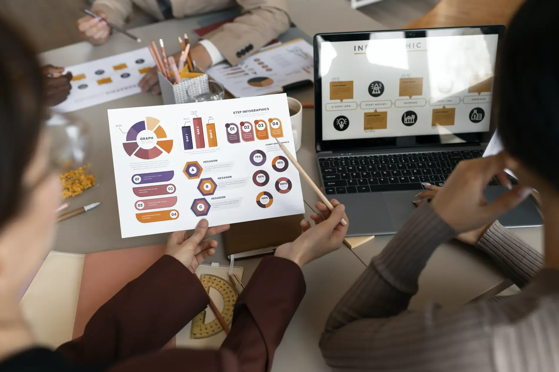 Persone a un tavolo di lavoro che analizzano grafici e infografiche stampati su un foglio, accanto a un laptop con altri grafici
