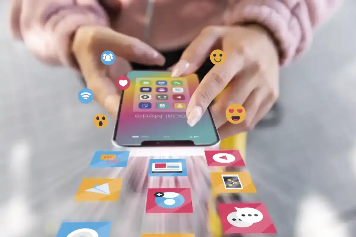 Mani che toccano uno smartphone da cui escono icone di app, emoji e notifiche dei social media